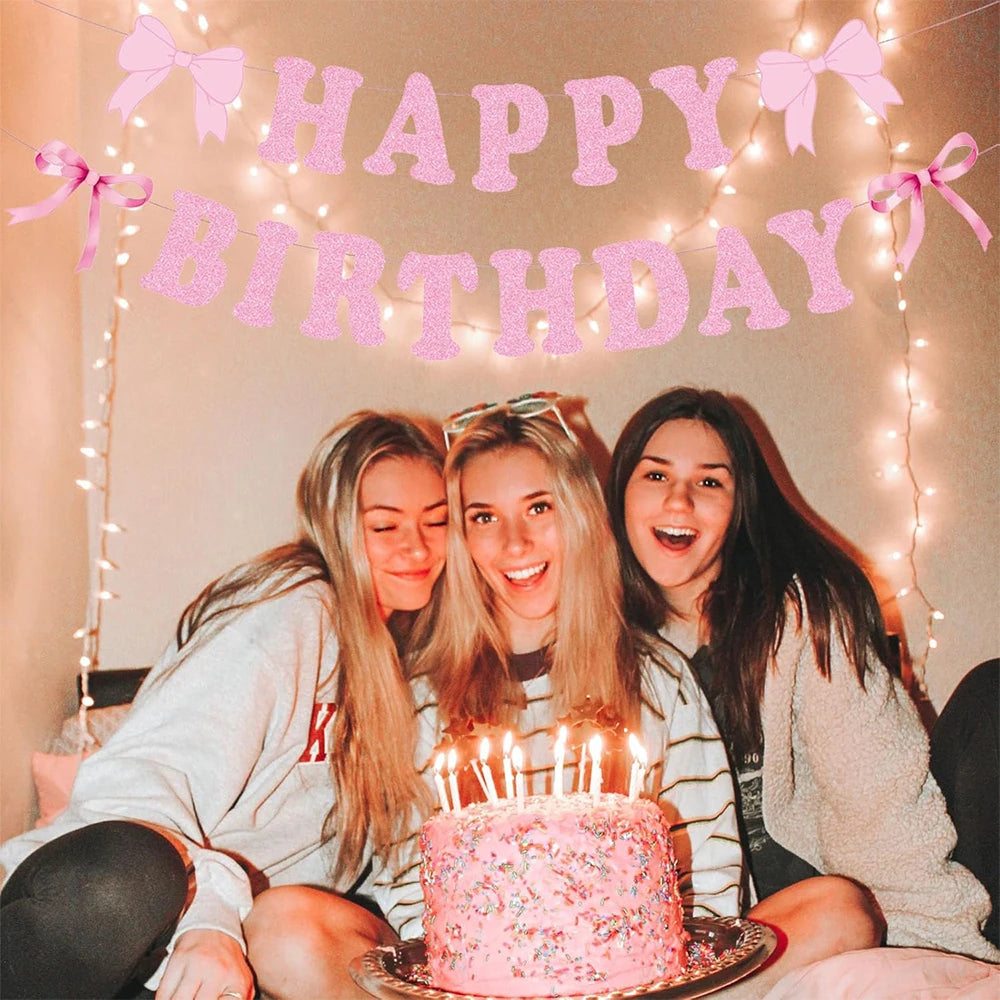 Three young women smiling while holding a glitter pink bow birthday banner in front of them.