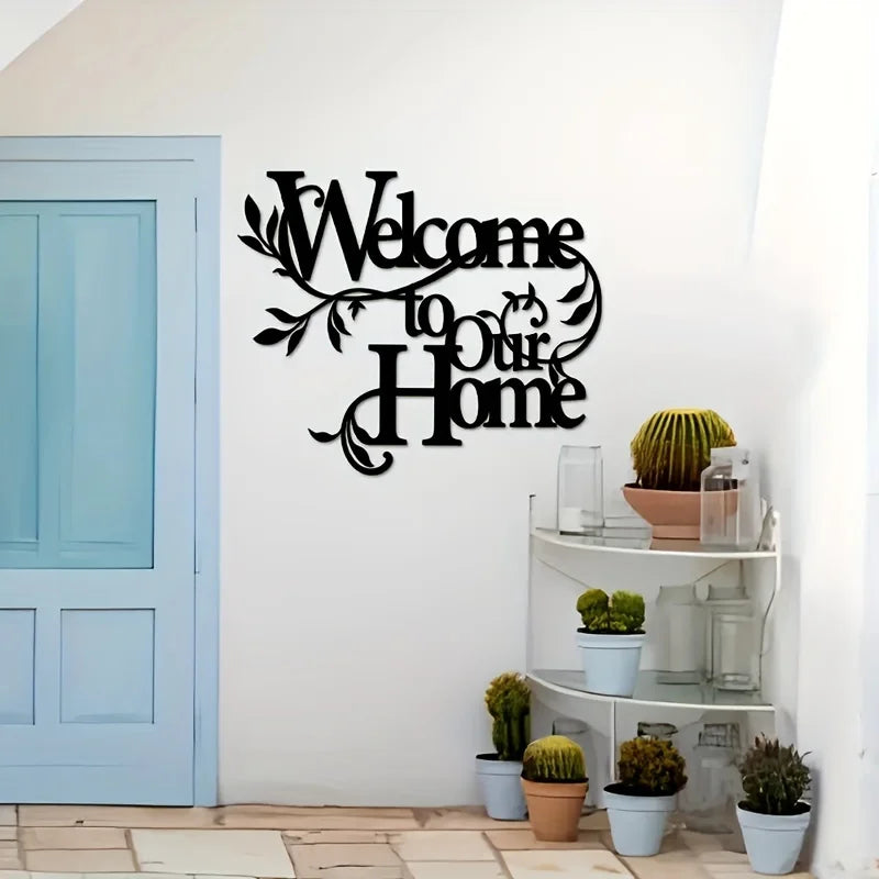A black metal welcome sign with cursive text on a white wall next to a light blue door and potted plants.