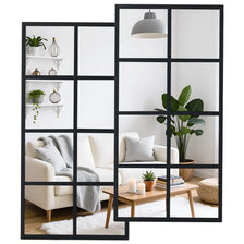 A modern window pane wall mirror with a black metal frame displaying interior decor and a sofa.