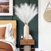 A tall vase of white artificial reed stems displayed on a wooden nightstand next to a bed.