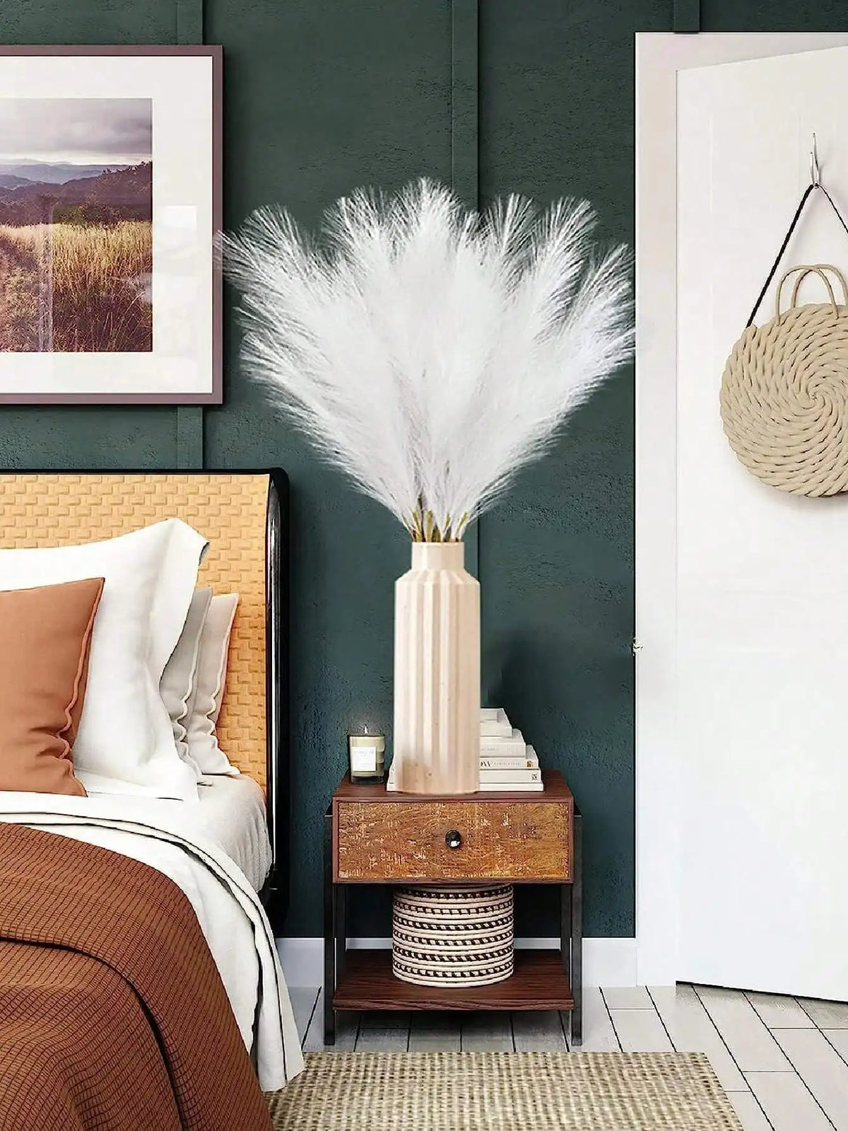 A tall vase of white artificial reed stems displayed on a wooden nightstand next to a bed.