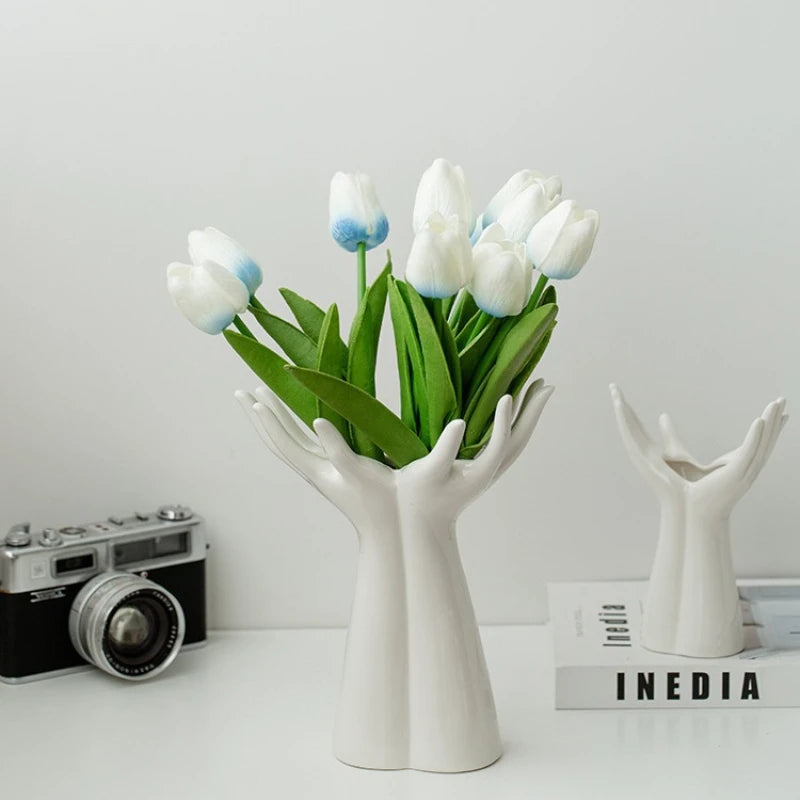 A white ceramic hand vase holds a bouquet of blue and white tulips on a white surface.