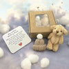 A beige teddy bear plush toy and an encouraging card are displayed next to a cardboard gift box against a soft purple backgro