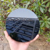 A black obsidian mirror with a geometric frame is held in a hand against a natural outdoor background.