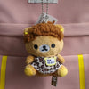 A brown Rilakkuma plush bear keychain hanging from a white tag against a pink fabric background.