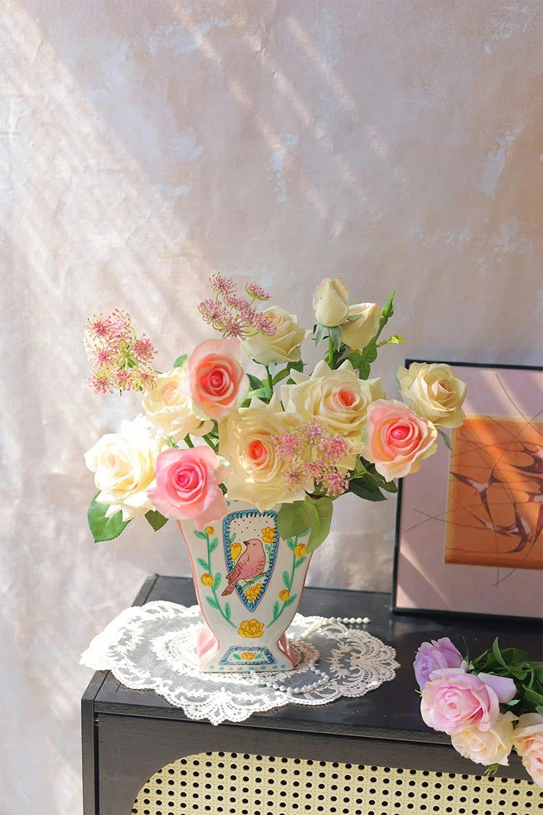 A bouquet of pink and cream roses and pink filler flowers in a ceramic bird floral vase on a dark table.