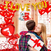 A couple embracing in front of a window with red heart balloons and a gold 'I love you' balloon decoration set.