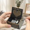 A hand holds an antique wood music box with engraved text and a small metal mechanism.