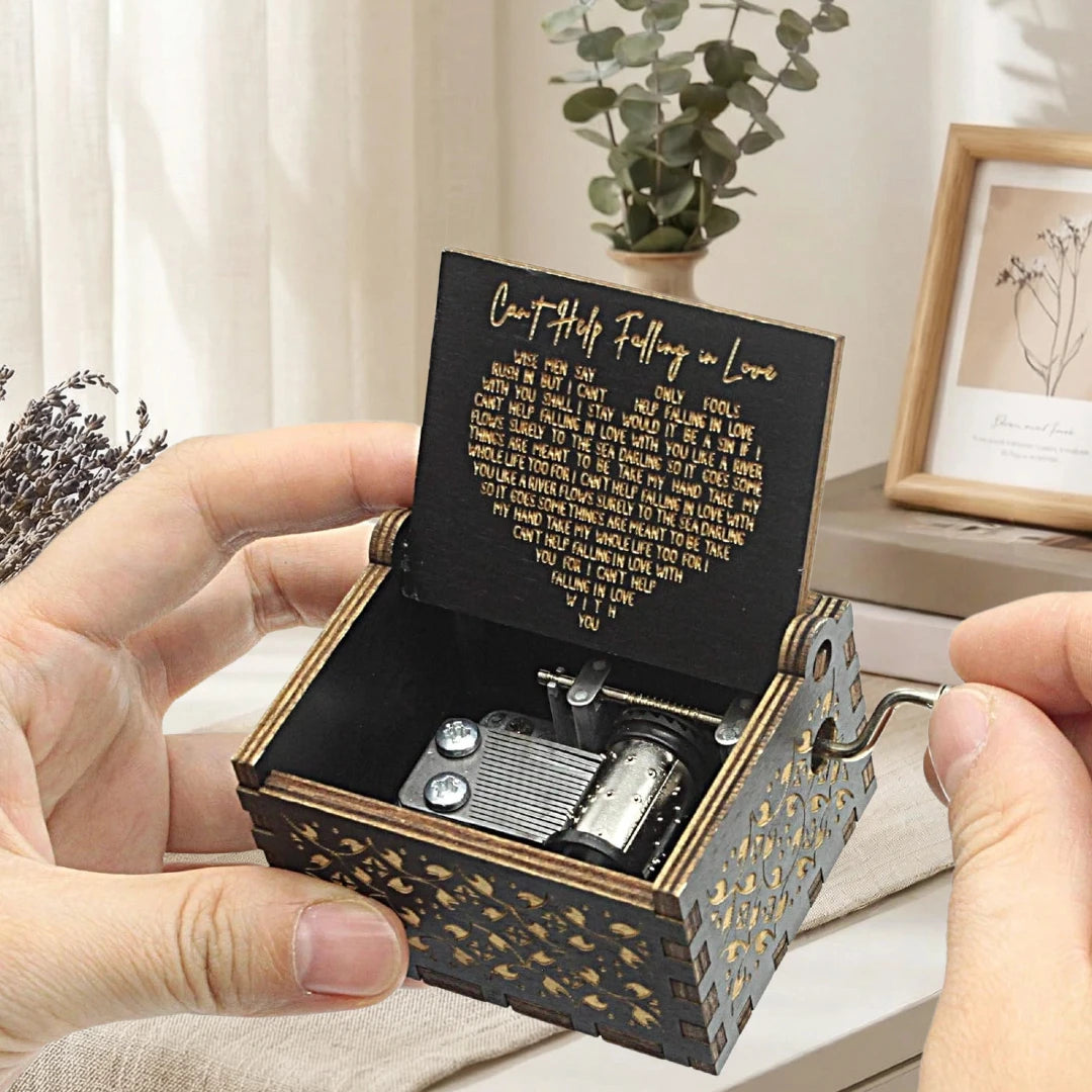A hand holds an antique wood music box with engraved text and a small metal mechanism.