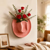 A pink ceramic wall vase holder with a rose arrangement hangs on a white wall next to a framed picture.