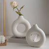 A white ceramic circular vase with a delicate rose stem is displayed on a light surface.