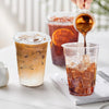 A hand pours dark coffee from a glass into a clear plastic cup filled with ice and liquid.