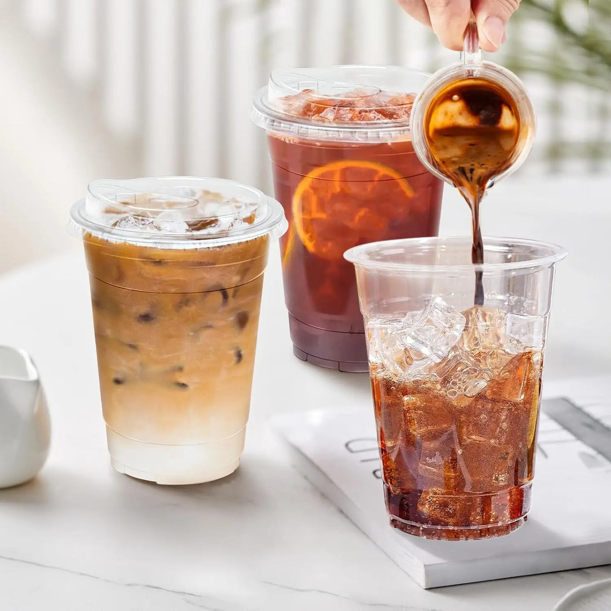 A hand pours dark coffee from a glass into a clear plastic cup filled with ice and liquid.