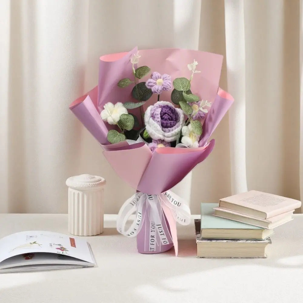 A pink handmade crochet rose flower bouquet with green leaves displayed on a white table.