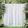 A white chiffon backdrop curtain decorated with floral arrangements stands between two wooden posts in a garden setting.