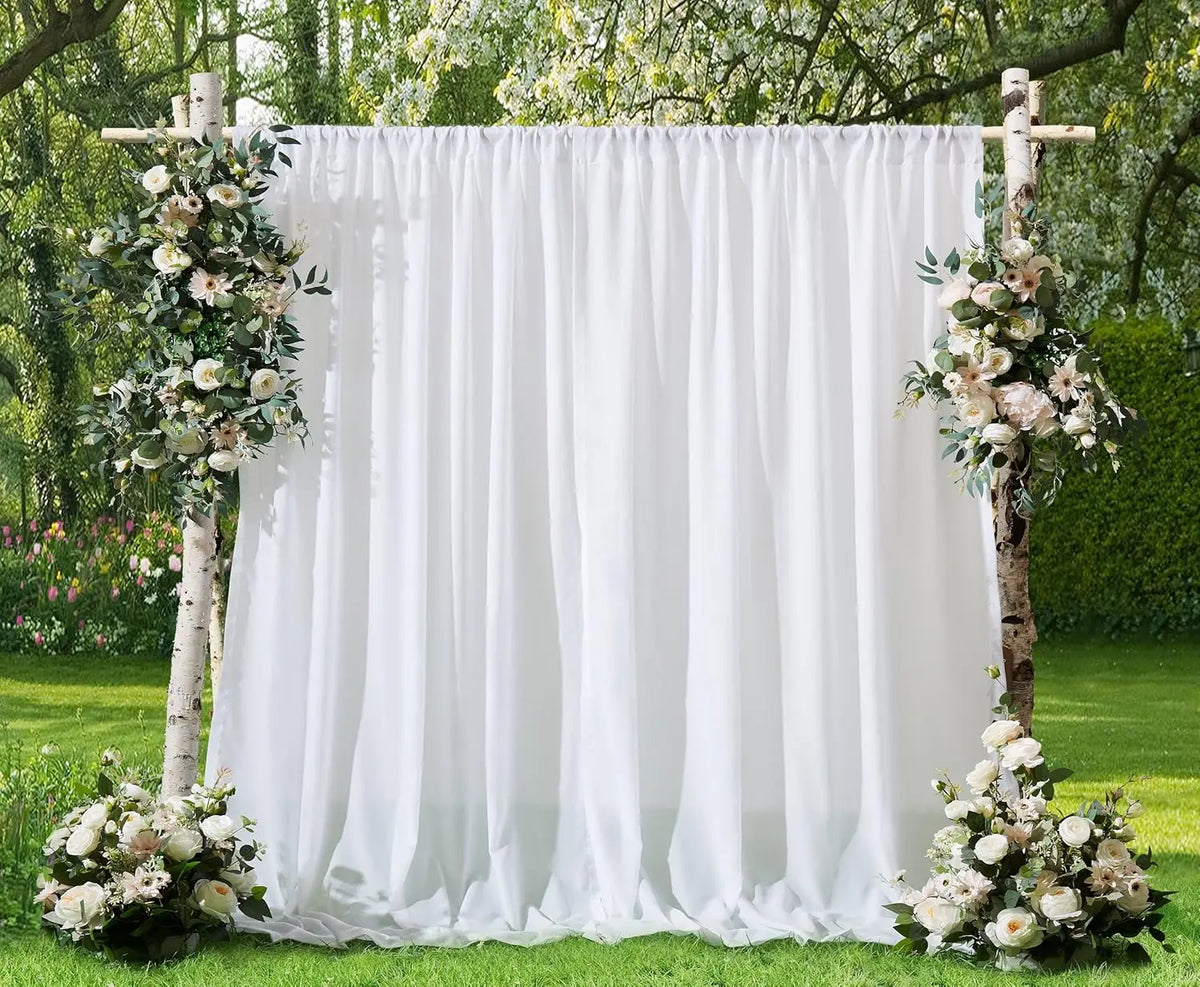 A white chiffon backdrop curtain decorated with floral arrangements stands between two wooden posts in a garden setting.