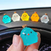 A hand holds a bright blue resin chicken car ornament next to several yellow and white chicken ornaments on a car dashboard.