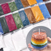 A collection of colorful sand pigments in small packets arranged in a row, with a finished candle candle in a glass jar nearb