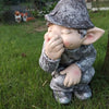 A ceramic statue of a gnome with closed eyes and a hand covering his mouth sitting in green grass.