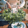 Une guirlande d'eucalyptus vert est placée sur une assiette blanche sur une table en bois, des mains l'arrangent.