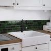 A white farmhouse sink is installed below a dark green subway tile backsplash in a kitchen setting.