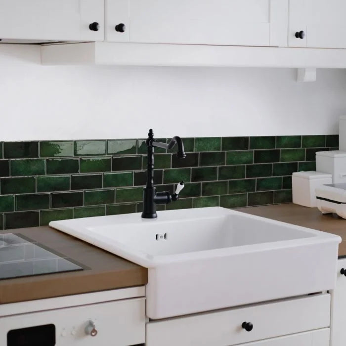 A white farmhouse sink is installed below a dark green subway tile backsplash in a kitchen setting.