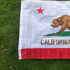 A white flag featuring a red star and a brown bear silhouette on the left, with the word CALIFORNIA in gold text across the b