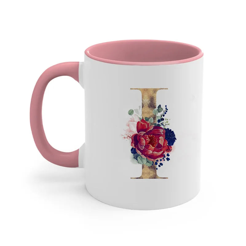 A pink ceramic coffee mug featuring a gold letter I decorated with colorful floral arrangements.
