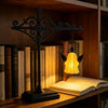 A black wrought iron lamp with a yellow glass shade hangs above an open book on a wooden shelf.