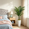 A potted artificial palm tree decor sits on a wooden nightstand next to a bed with light blue bedding.