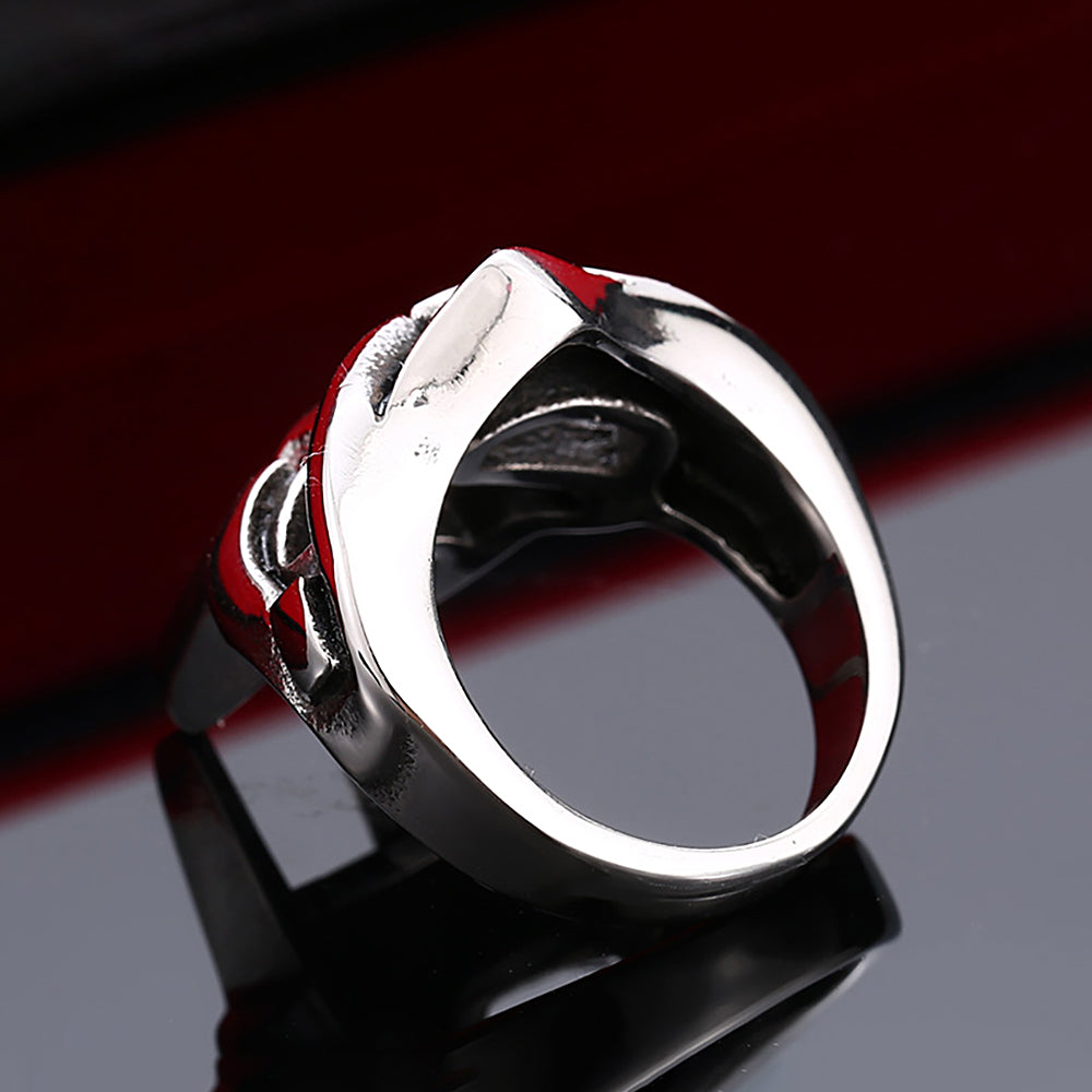 A silver stainless steel ring featuring a mask design with red accents, displayed on a dark reflective surface.