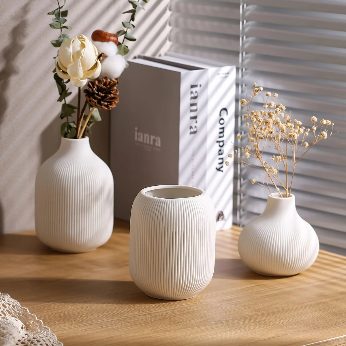 Three minimalist white ceramic decorative vases with dried flowers and natural elements on a wooden surface.