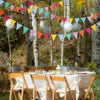 A string of colorful pennant banners hangs between birch trees in an outdoor setting.