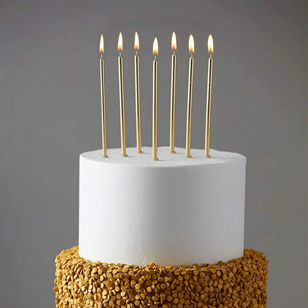 A white birthday cake topped with thirty metallic gold candles in holders, resting on a layer of gold-colored cereal.
