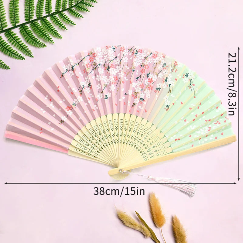 A pink and green elegant bamboo folding hand fan with a tassel displayed on a light purple background.