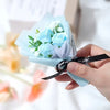 Une main tient un bouquet de mini roses artificielles bleu clair avec des fleurs de remplissage blanches et un ruban noir.