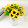 A bouquet of bright yellow artificial sunflowers in a clear glass vase on a white surface.