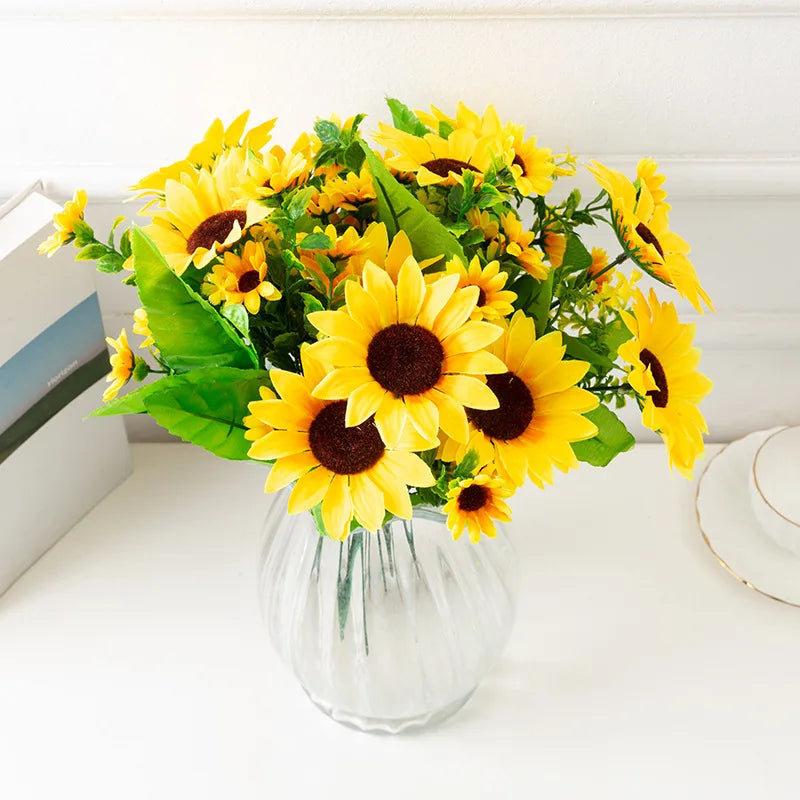 A bouquet of bright yellow artificial sunflowers in a clear glass vase on a white surface.