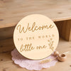 A round wooden welcome sign with gold lettering that reads Welcome to the world little one, resting on pink lace.