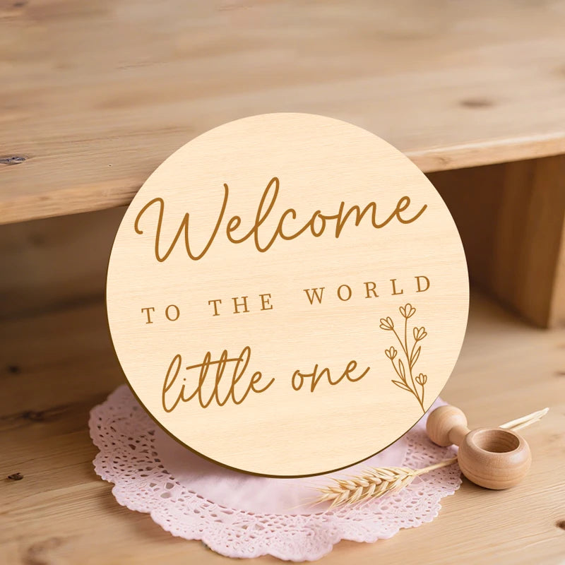 A round wooden welcome sign with gold lettering that reads Welcome to the world little one, resting on pink lace.