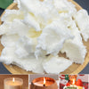 A pile of white natural coconut wax pieces on a wooden board, next to candles and a coconut candle.