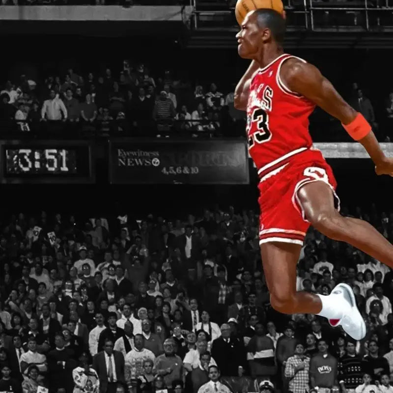 A male basketball player in a red jersey is captured mid-air during a slam dunk, with a crowd visible in the background.