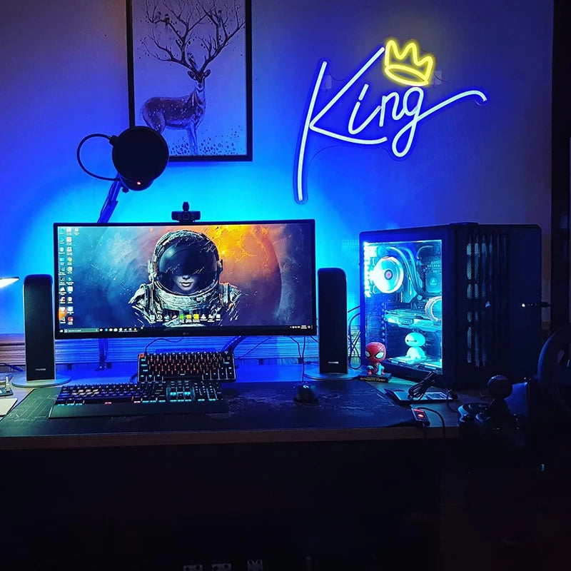 A desk setup featuring a computer monitor displaying a space scene, surrounded by neon lighting and a crown sign.