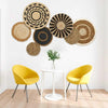 A boho straw wall hanging with woven tassels is displayed above a white table and two yellow chairs.