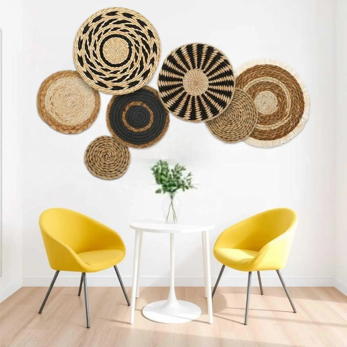 A boho straw wall hanging with woven tassels is displayed above a white table and two yellow chairs.