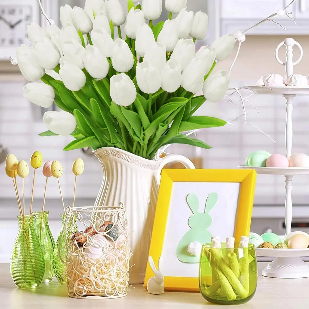 Un ramo de tulipanes de PU blancos artificiales en un jarrón rodeado de pequeños objetos decorativos y follaje verde.