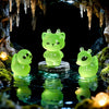 A glowing green resin cat and duck charm resting on a dark surface surrounded by moss and cave formations.