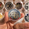 A hand holds a silver-plated commemorative coin featuring a cross design and the scripture Ephesians 6:11.
