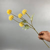 Una mano sostiene un tallo de flores artificiales de margarita/diente de león amarillo contra un fondo gris.