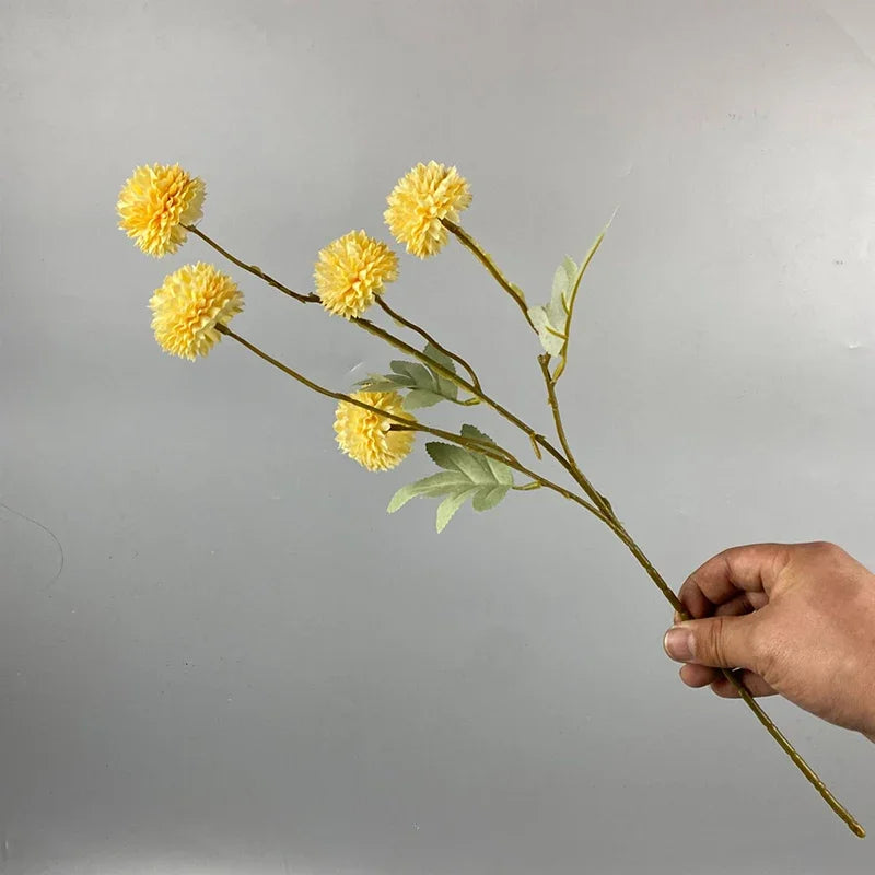 Una mano sostiene un tallo de flores artificiales de margarita/diente de león amarillo contra un fondo gris.